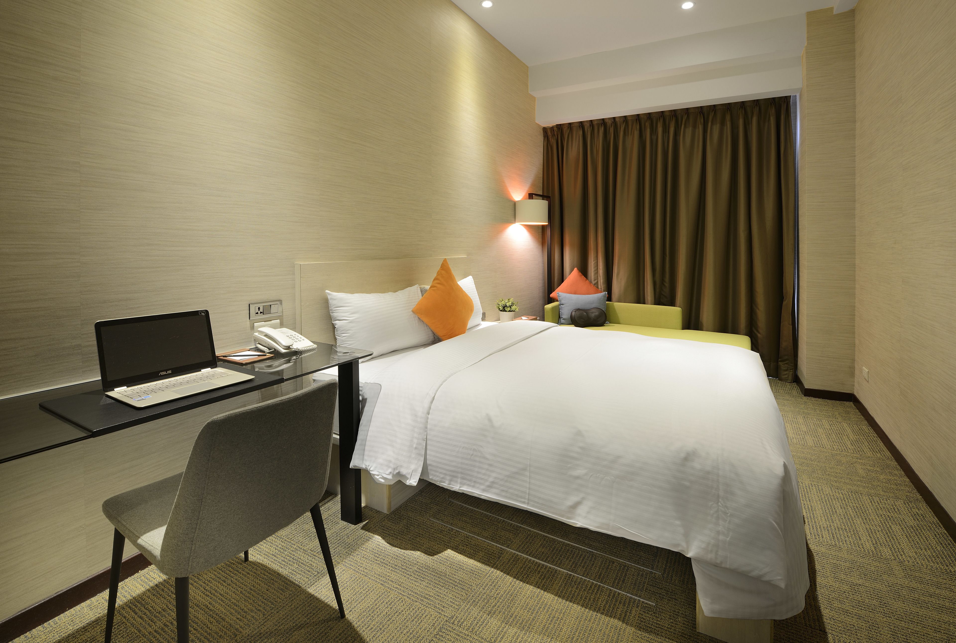 A hotel room with a bed, desk, laptop, chair, and a lamp.