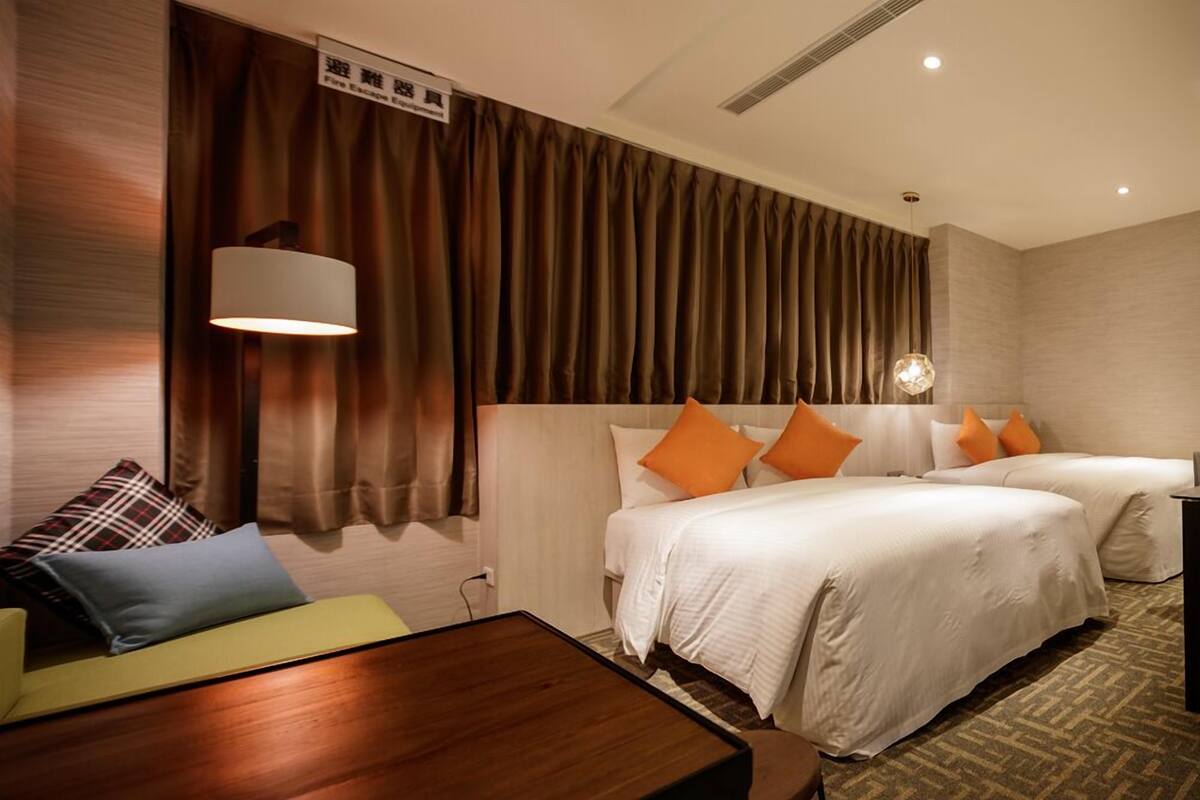 A hotel room with two beds, a sofa, a wooden side table, and a large window with curtains.