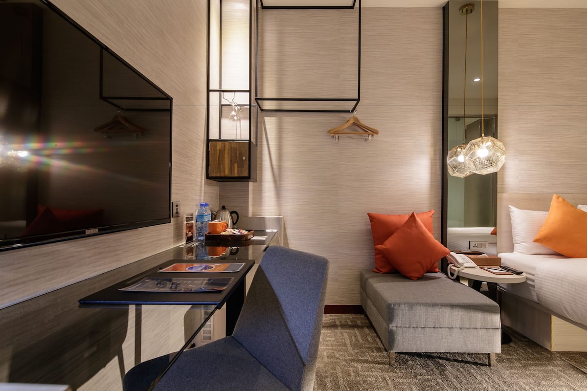 A modern hotel room with a large flat-screen TV, a sofa, a small table, and a bed with orange pillows.