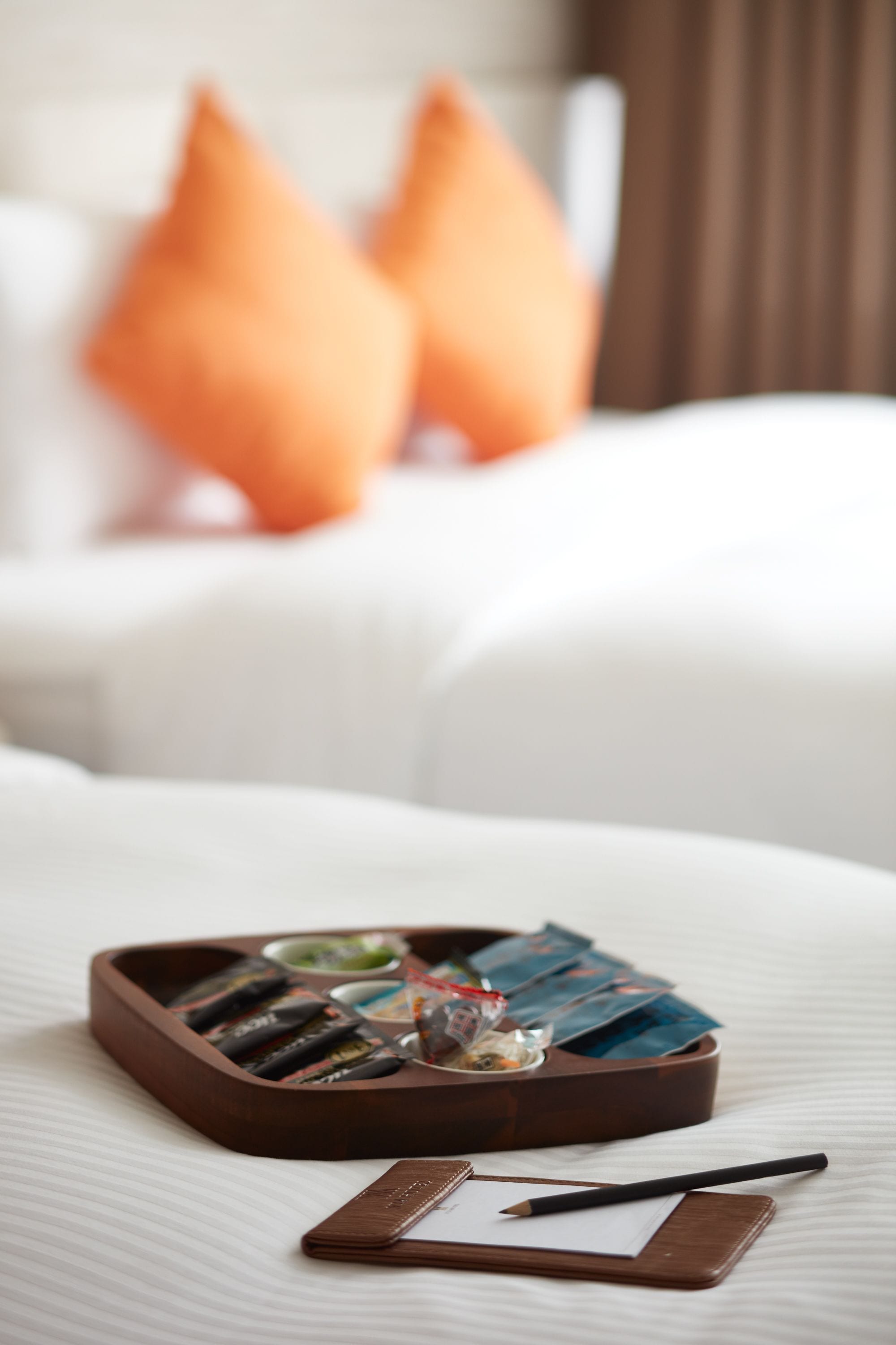 Bandeja bem organizada com chocolates e um pequeno copo sobre uma cama com roupa de cama branca e almofadas laranja.