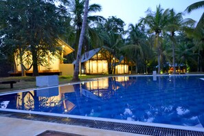 A swimming pool with a clear reflection, surrounded by palm trees and a building with warm lighting.