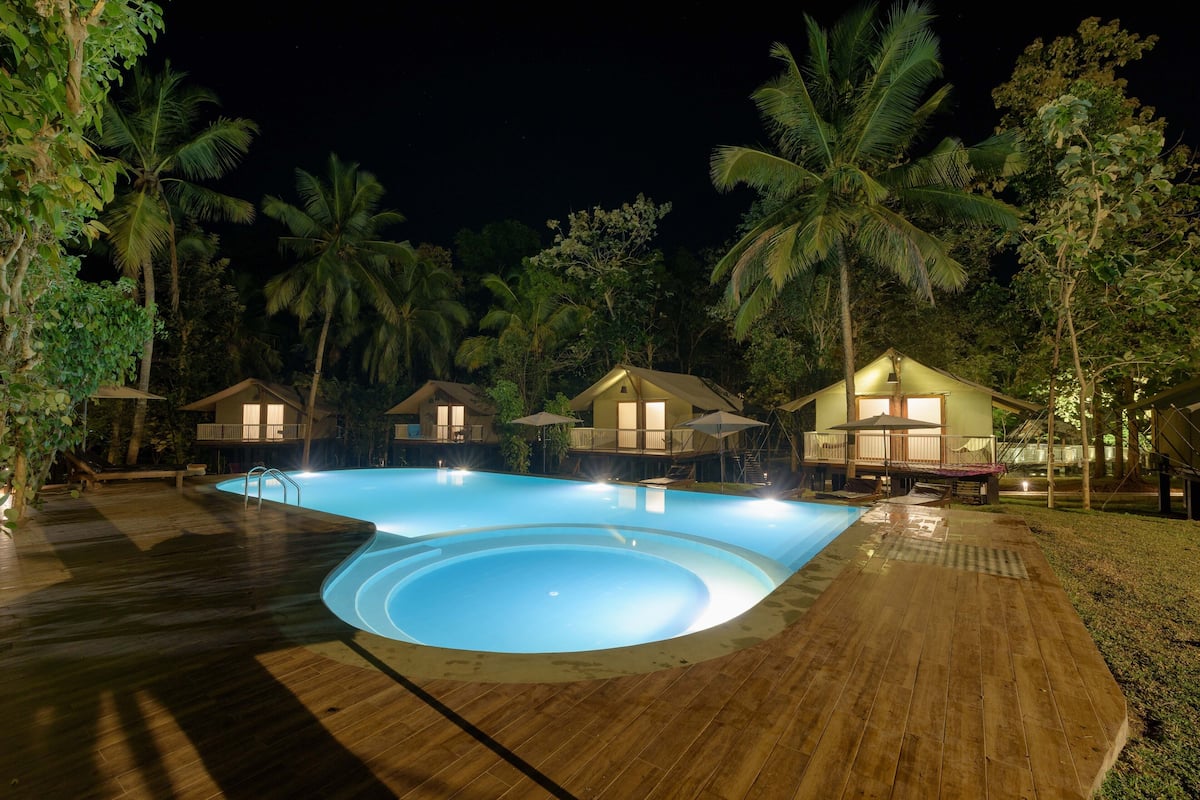 Una piscina rodeada de terrazas de madera y palmeras, con varias casas pequeñas al fondo.