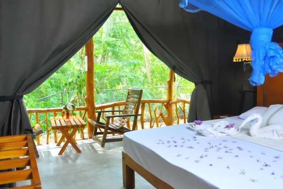 Una habitación con cama, una silla de madera, una mesita y un balcón con vistas a la vegetación.