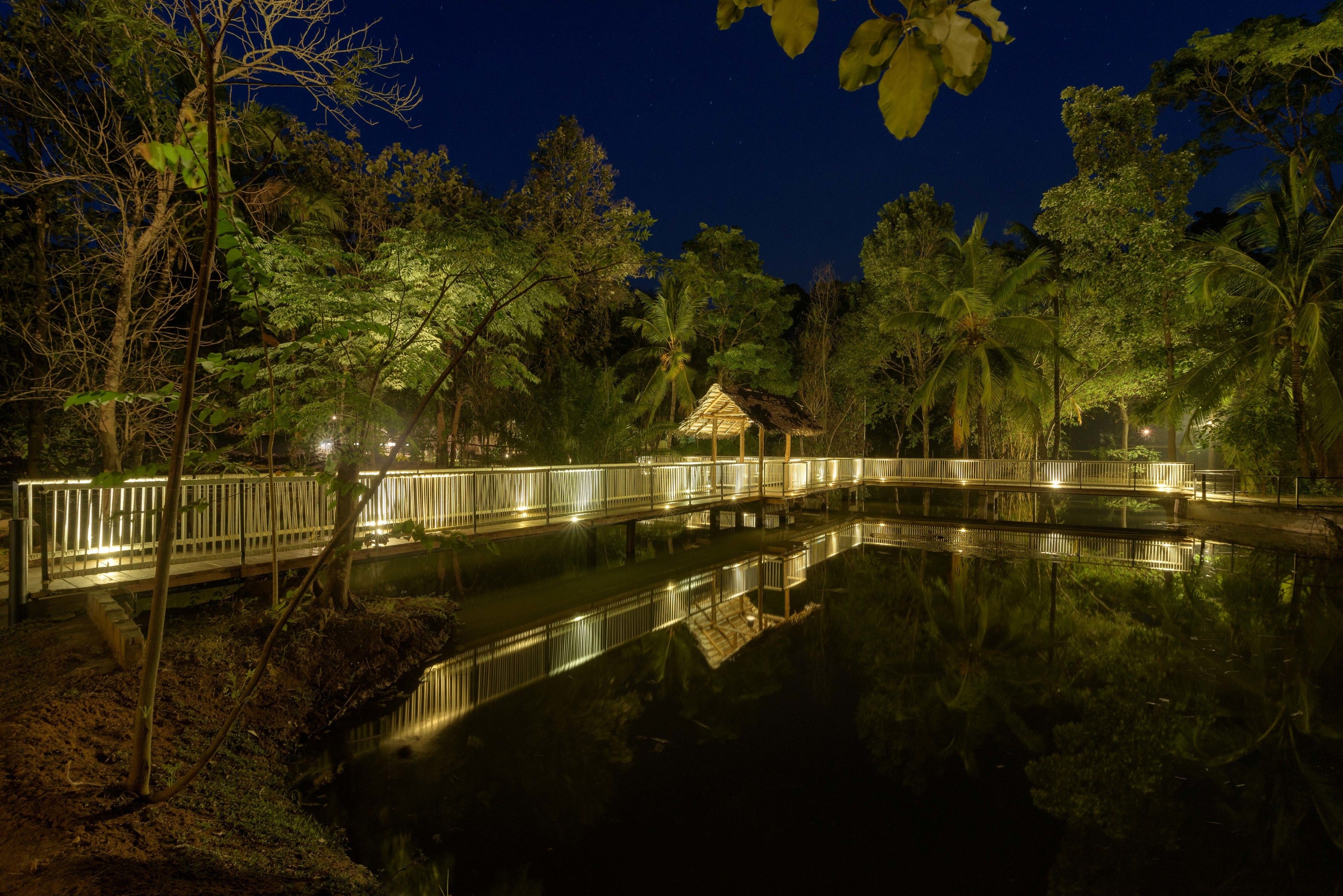 Vista nocturna de un estanque con un puente de madera y una cabaña con techo de paja, rodeada de árboles.