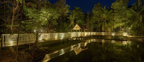 Vista nocturna de un estanque con un puente de madera y una cabaña con techo de paja, rodeada de árboles.