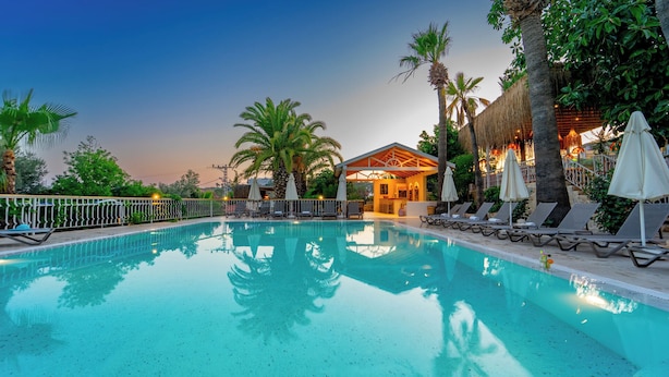 A pool area with lounge chairs and umbrellas, surrounded by palm trees and a building with a thatched roof. - Golden Lighthouse Hotel (Kas)