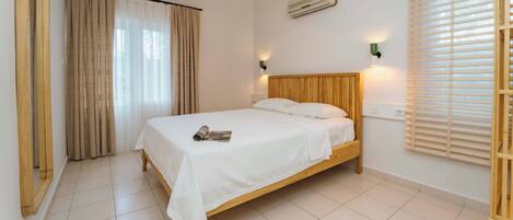 Un dormitorio con cama de madera, ropa de cama blanca, una ventana con persianas, aire acondicionado y una lámpara en la pared.