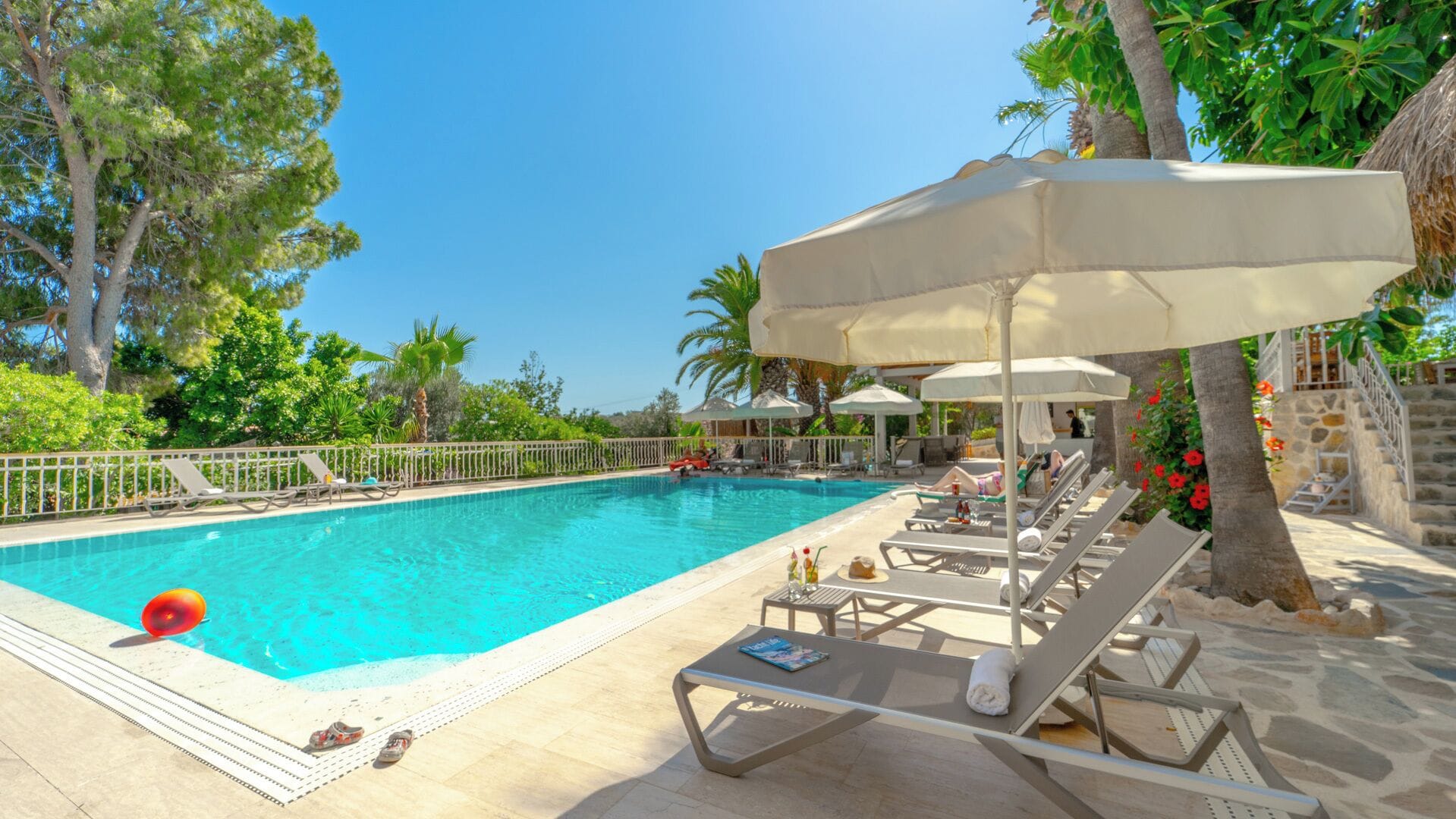 A pool area with lounge chairs and umbrellas, surrounded by trees and a stone wall.