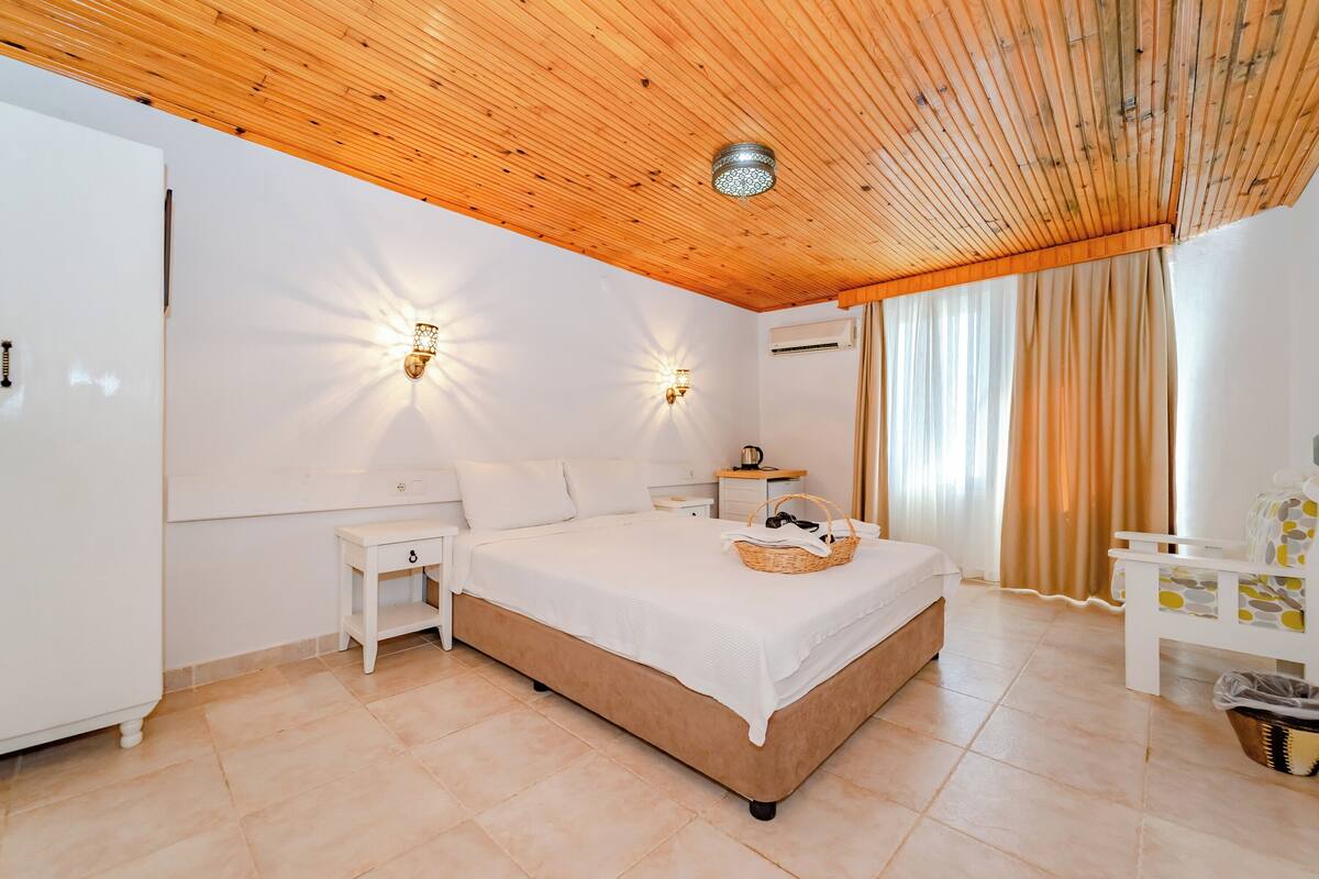 A bedroom with a wooden ceiling, a bed with a white bedspread, two wall-mounted lights, a nightstand, and a window with curtains.
