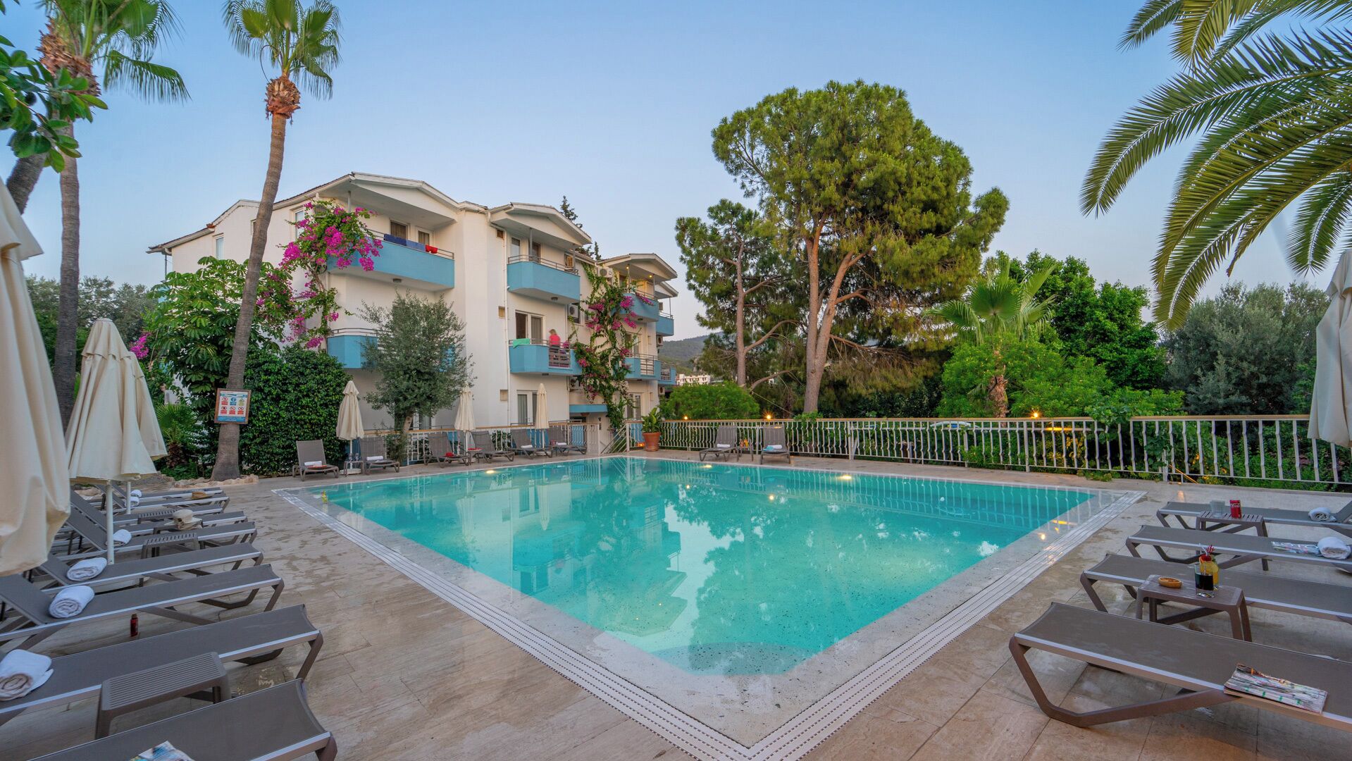 A swimming pool with lounge chairs and umbrellas, surrounded by a building with balconies and trees.