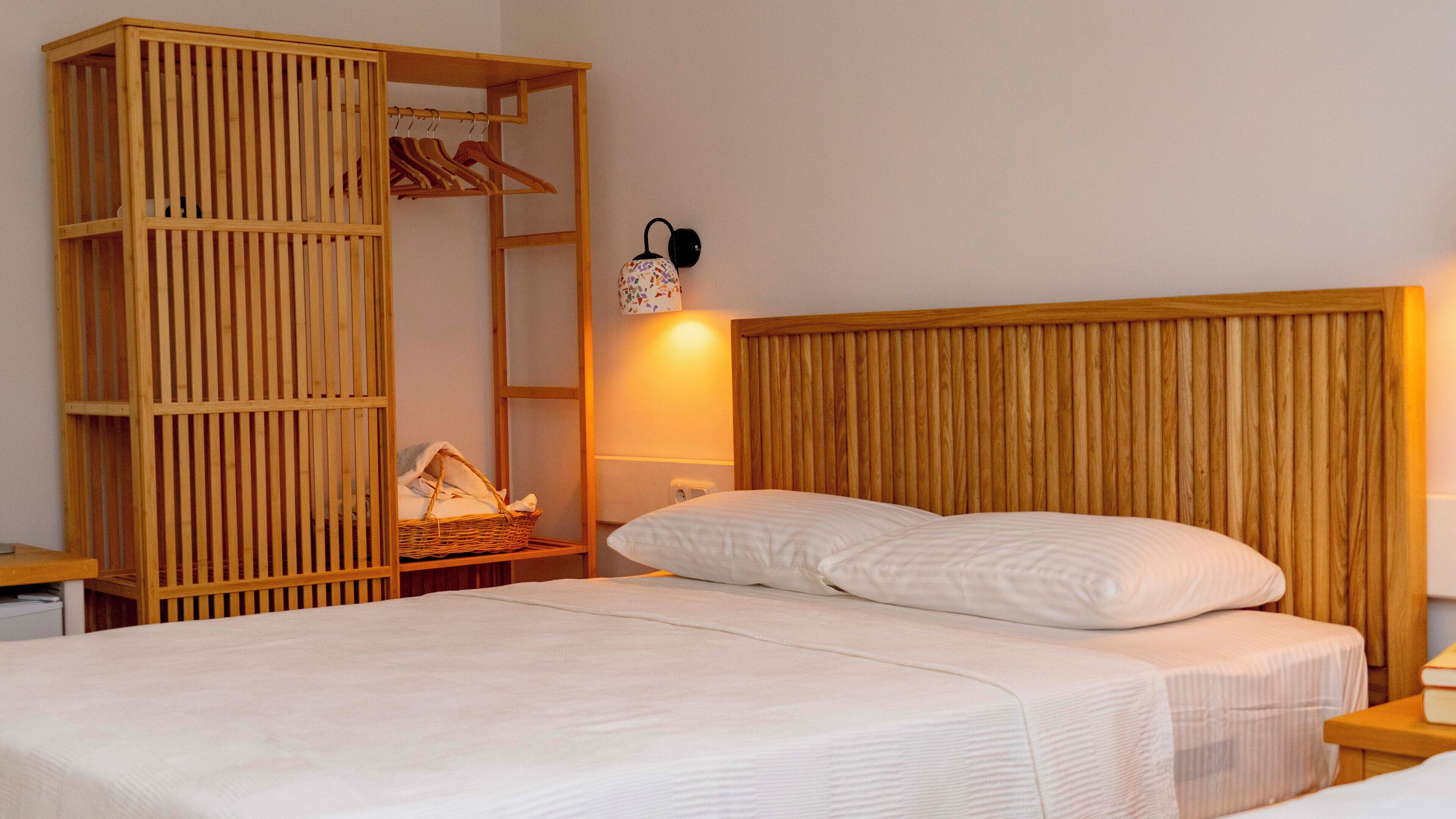 A bedroom with a wooden headboard, a bed with white linens, a wooden wardrobe with clothes and a basket, and a wall-mounted lamp.
