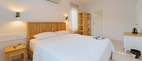 Un dormitorio con cabecera de madera, una mesita de noche con control remoto, un armario de madera, una puerta blanca y una pequeña cocina con hervidor.