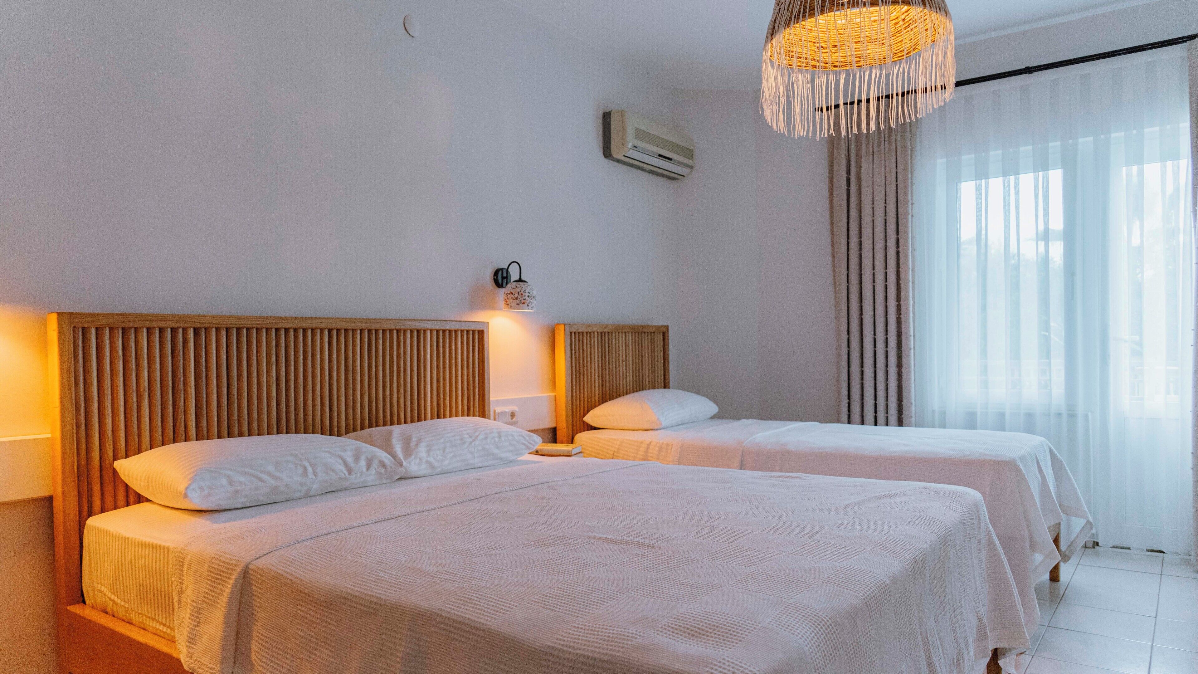 A hotel room with two beds, a wooden headboard, a wall-mounted air conditioner, a hanging light fixture, and a window with sheer curtains.