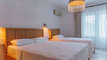 A hotel room with two beds, a wooden headboard, a wall-mounted air conditioner, a hanging light fixture, and a window with sheer curtains.