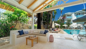 A poolside area with a sofa, table, and chairs, surrounded by tropical plants and a clear blue sky. - Golden Lighthouse Hotel (Kas)