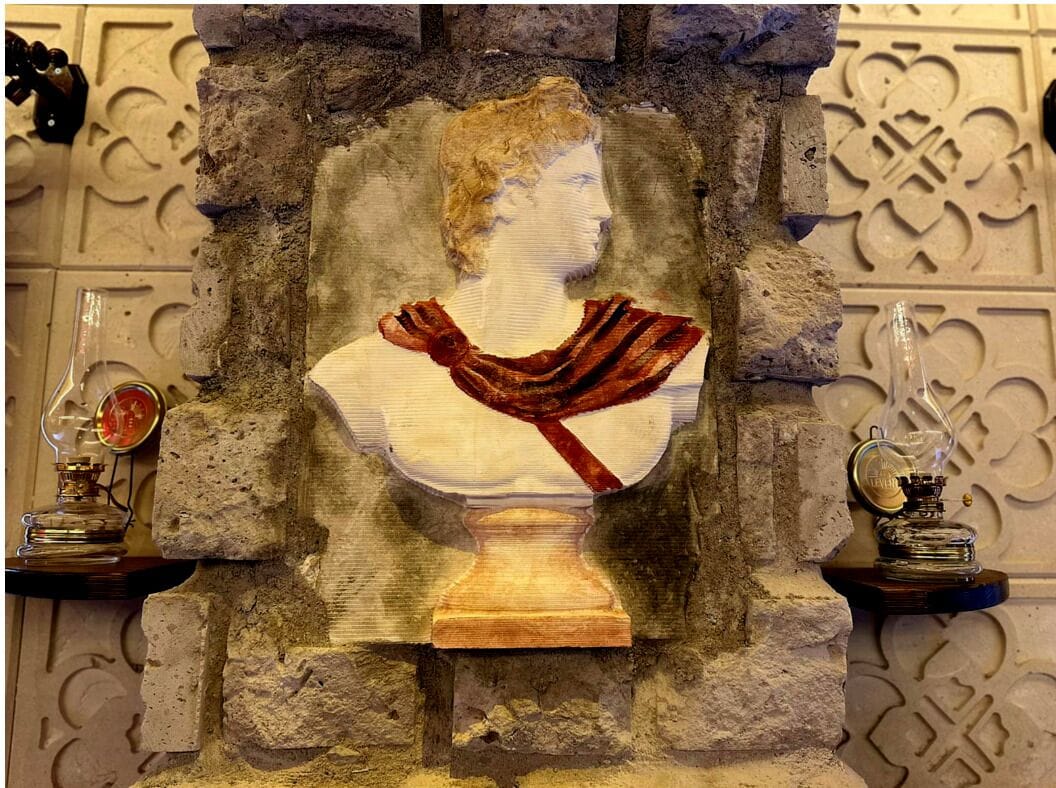 Escultura em busto com um tecido vermelho drapeado, em frente a uma parede de pedra texturizada com padrões decorativos.