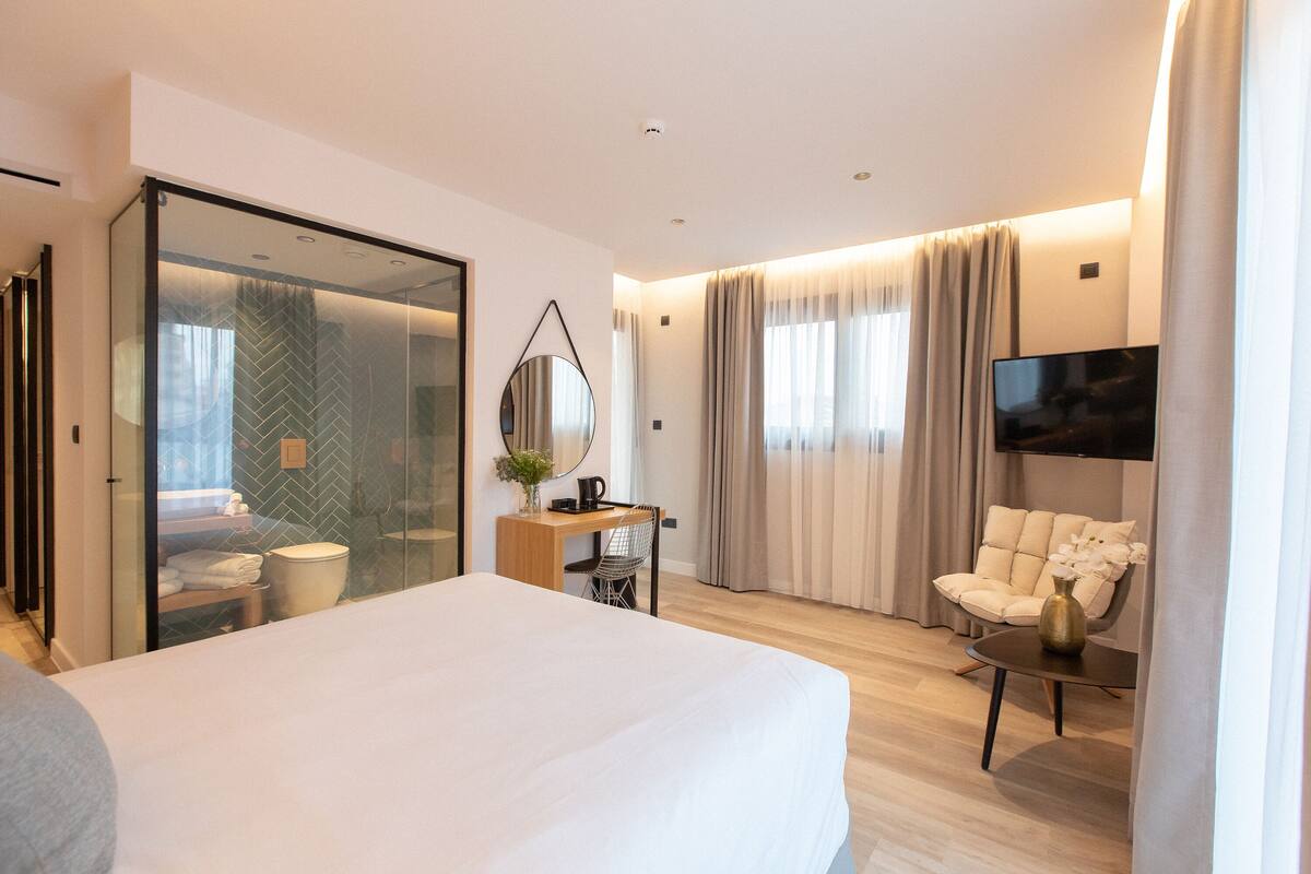 Una habitación de hotel moderna con una cama grande, un escritorio con una silla, un televisor y una pared divisoria de vidrio.