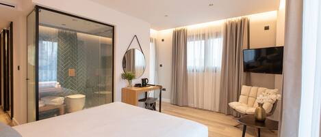 A modern hotel room with a large bed, a desk with a chair, a TV, and a glass partition.