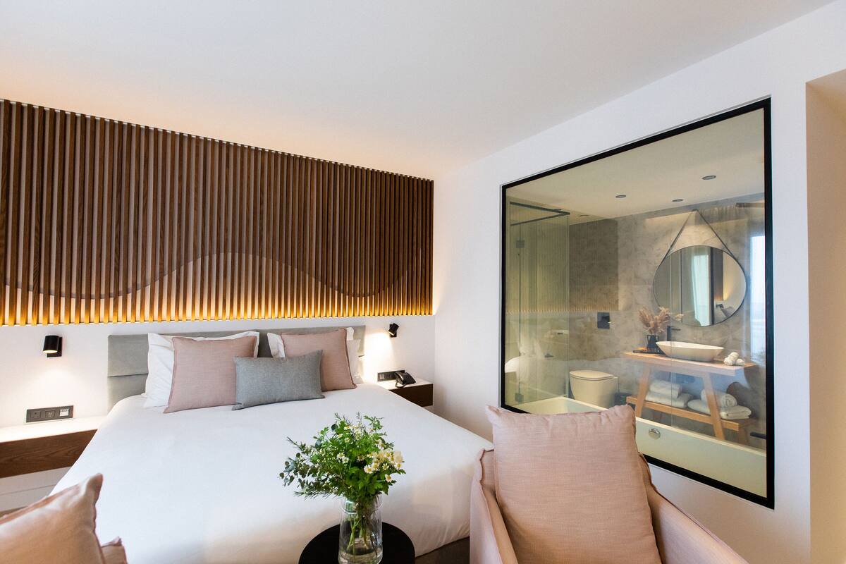 Una habitación de hotel moderna con una cama grande, una mesita de noche, un tocador y vistas al baño.