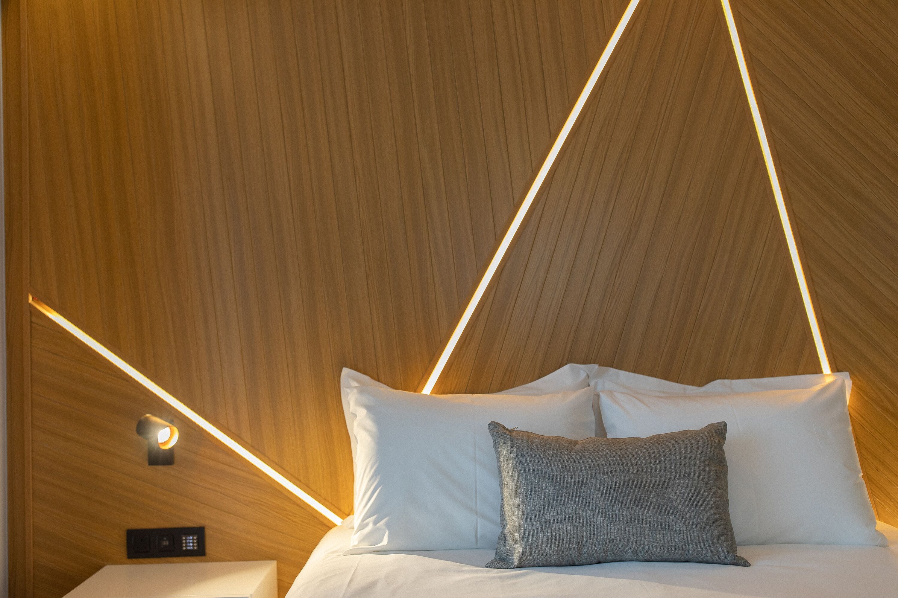 A modern hotel room with a wooden ceiling, a bed with white and gray pillows, and a control panel on the wall.