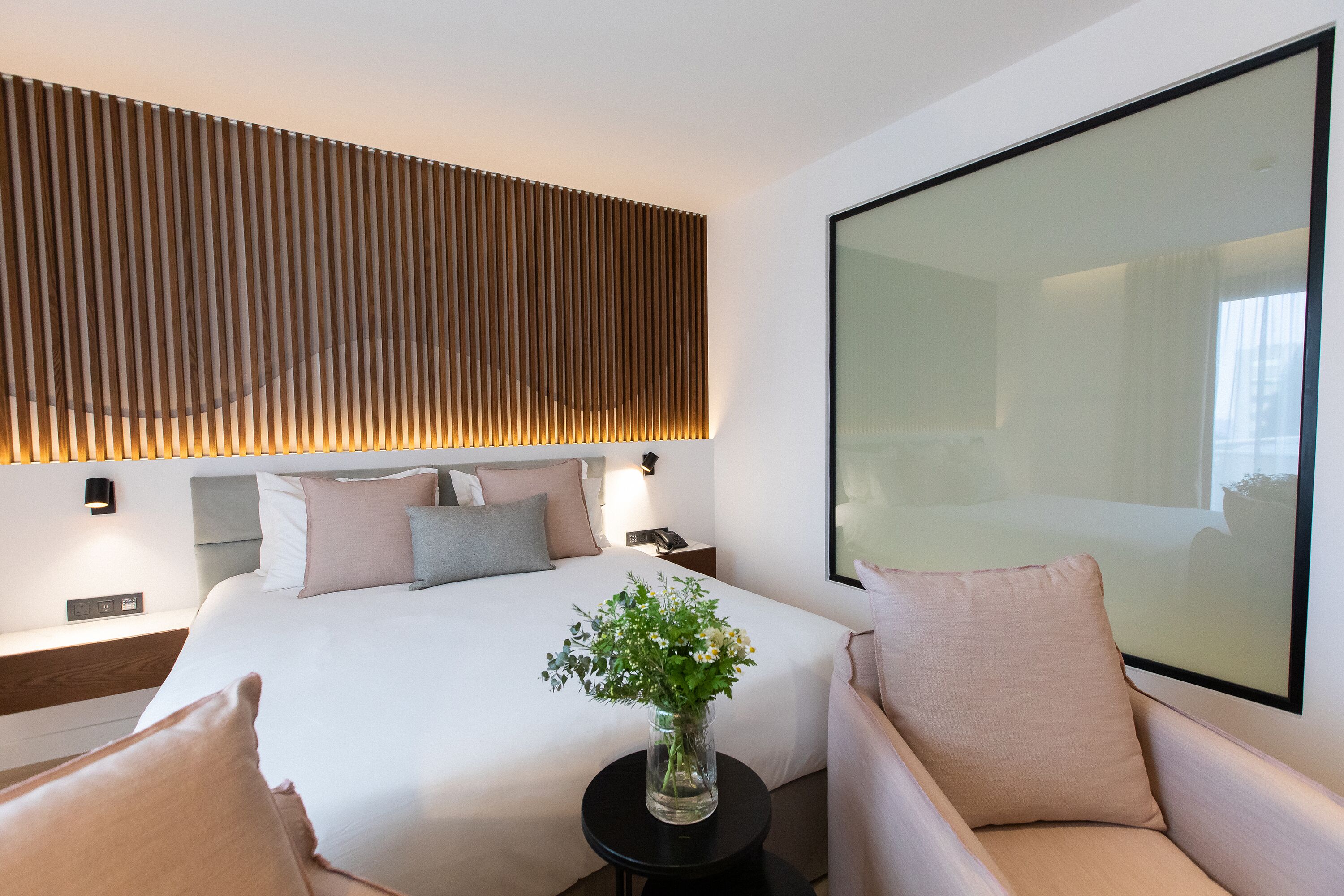 Una habitación de hotel moderna con una cama grande, una mesita de noche, una pequeña mesa redonda con un jarrón de flores y un espejo grande en la pared.