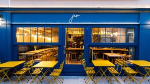 Exterior azul de un restaurante con mesas y sillas amarillas, ventanas grandes y un interior visible con mesas y sillas.