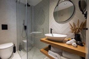 A modern bathroom with a glass shower enclosure, a white sink with a vessel basin, and a wooden vanity with towels and toiletries. - Old Port Hotel (Limassol)