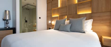 A modern hotel room with a large bed, wooden headboard with integrated lighting, a bedside table, and a glass shower enclosure.