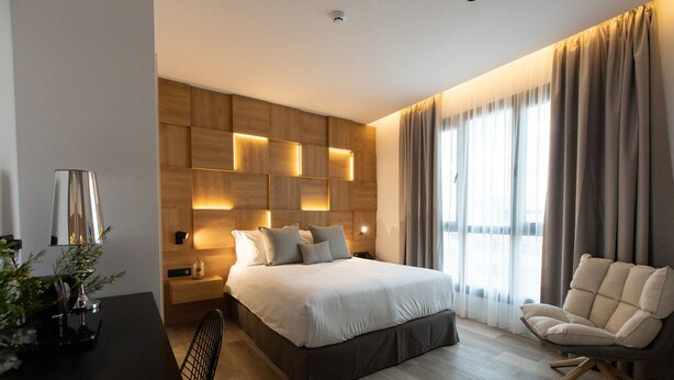 Un dormitorio moderno con una cama grande, una mesita de noche con lámpara, una silla y un cabecero de madera con iluminación integrada.