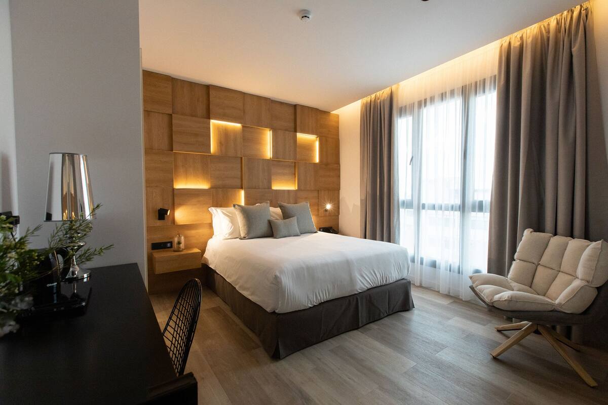 Un dormitorio moderno con una cama grande, una mesita de noche con lámpara, una silla y un cabecero de madera con iluminación integrada.