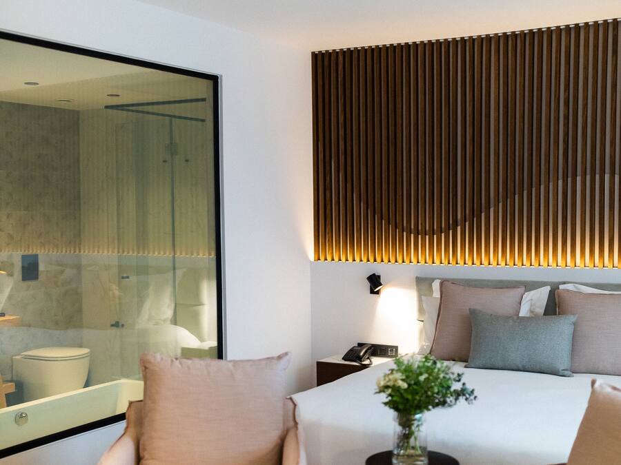 A modern hotel room with a large bed, two armchairs, a small table with a vase, and a large mirror.