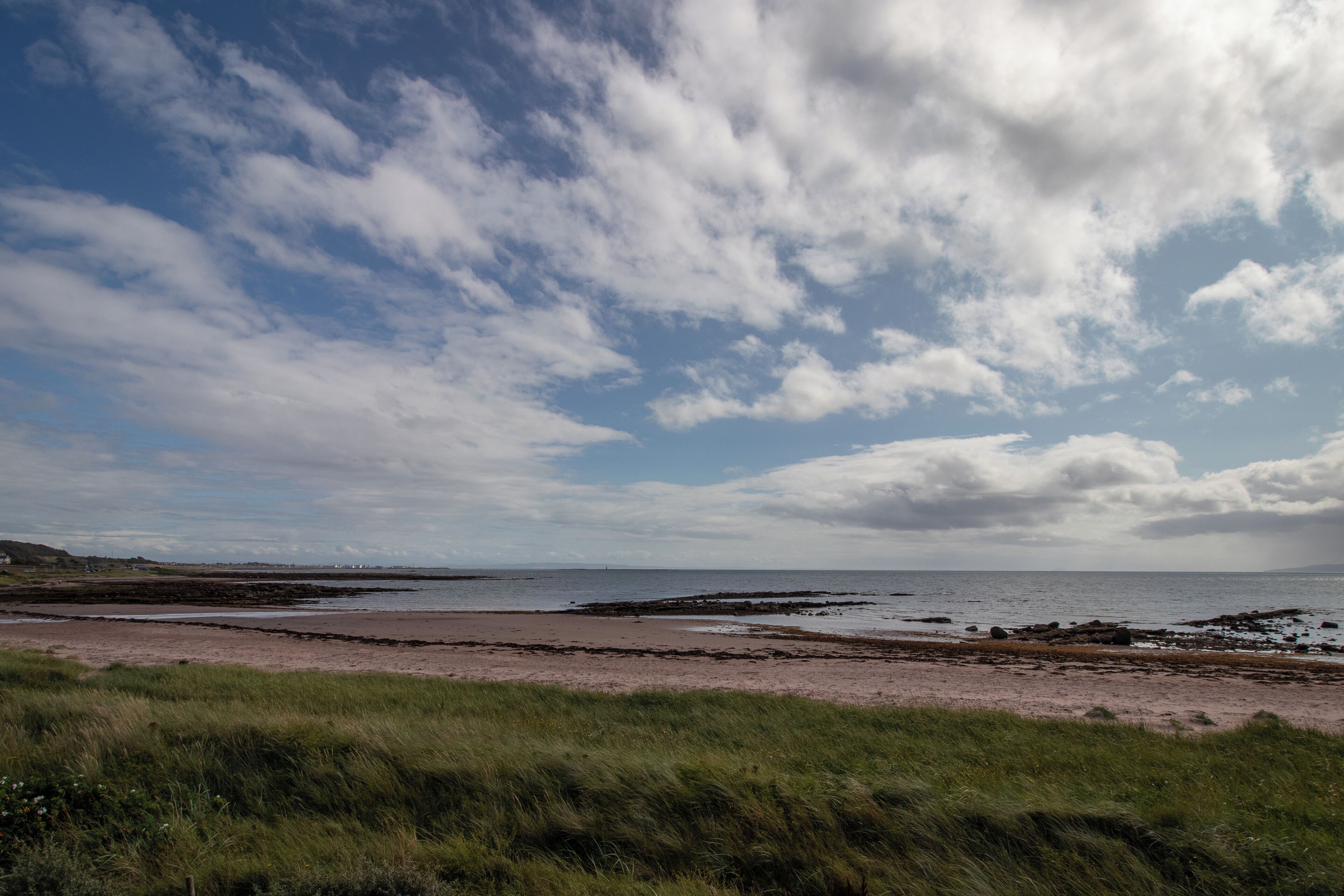 A coastal landscape with a beach, grassy foreground, and a partly cloudy sky.