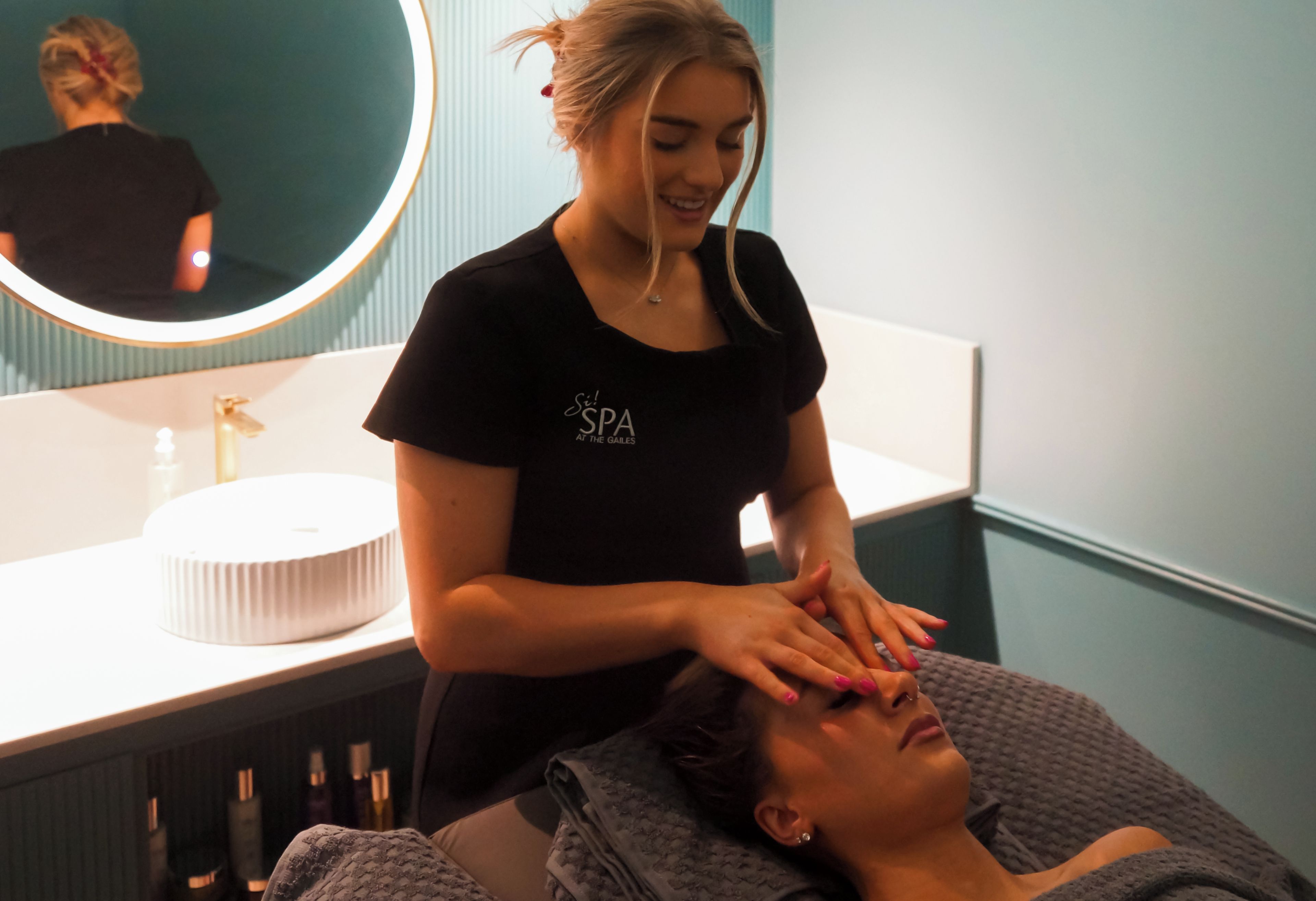 A person receiving a facial treatment in a spa setting with a mirror and a sink in the background.