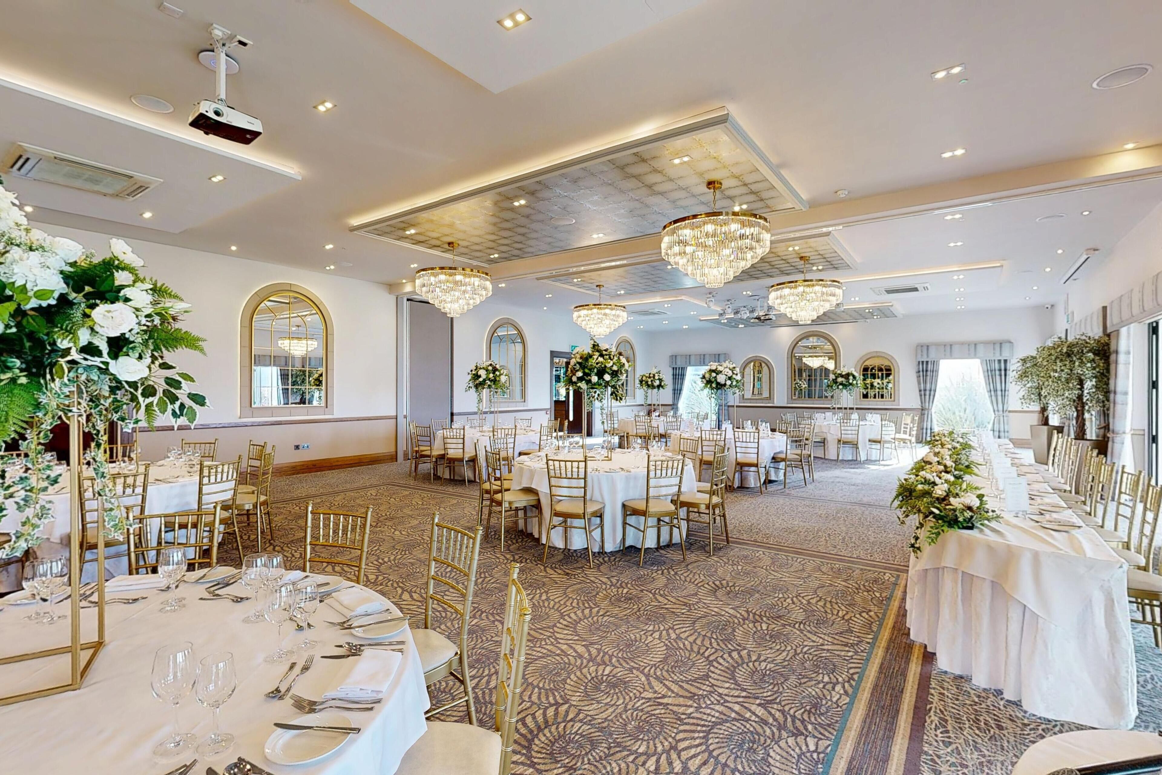 Un salón de banquetes con mesas redondas, sillas doradas, candelabros y centros de mesa florales.