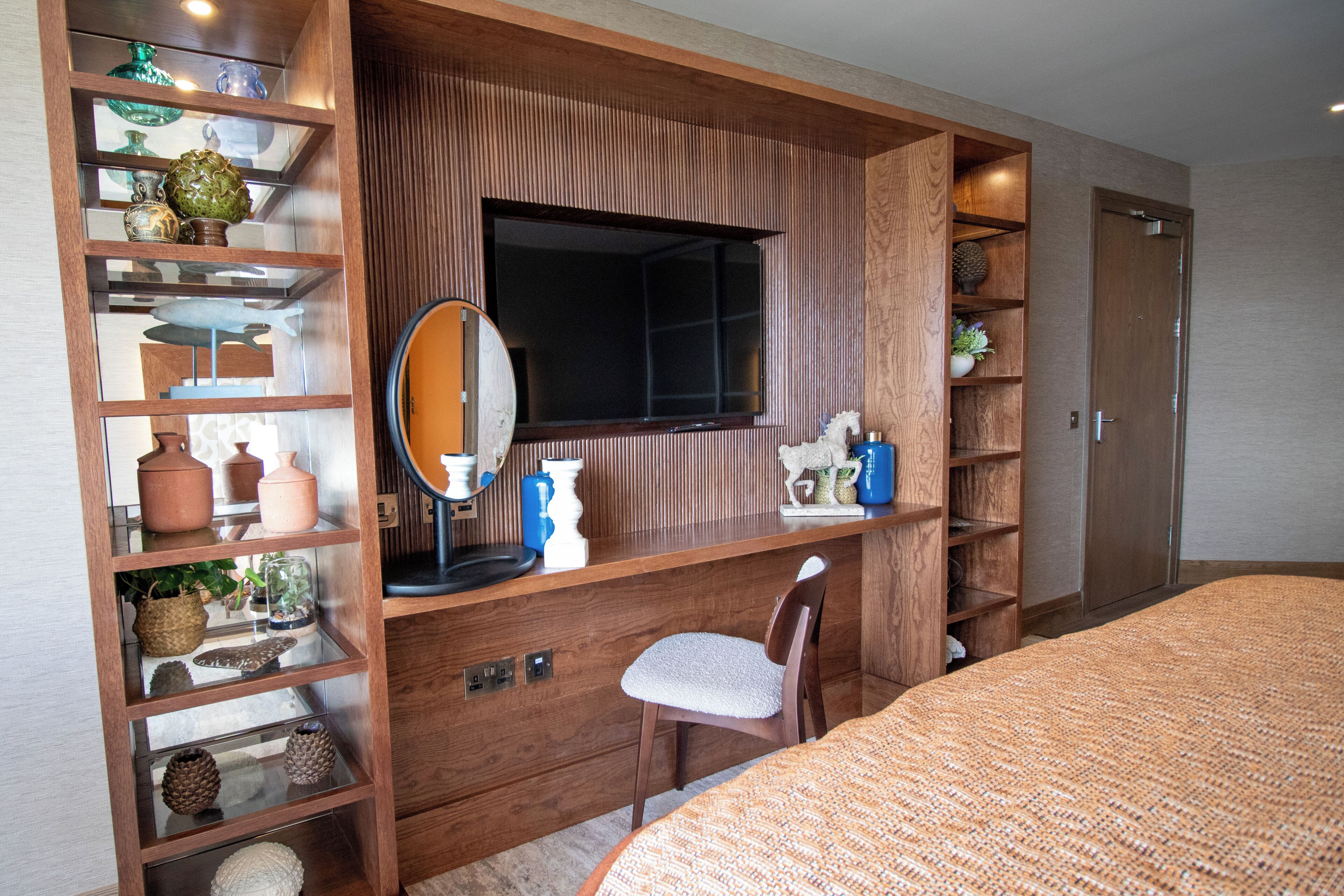A bedroom with a bed, a wooden desk with a mirror, shelves with decorative items, and a wardrobe.