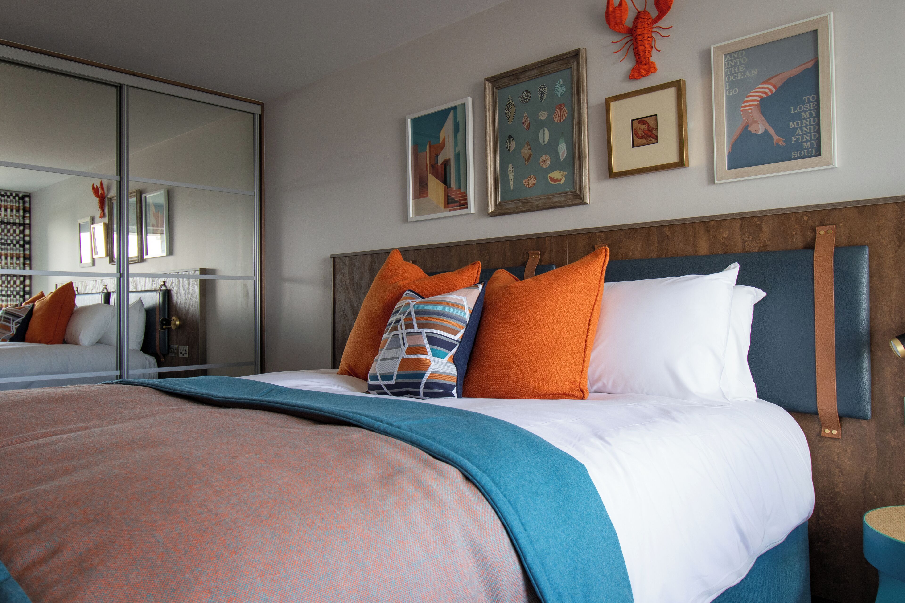 A modern hotel room with a large bed, orange and white pillows, a blue headboard, and artwork on the walls.