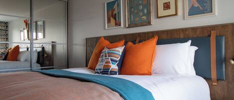 A modern hotel room with a large bed, orange and white pillows, a blue headboard, and artwork on the walls.