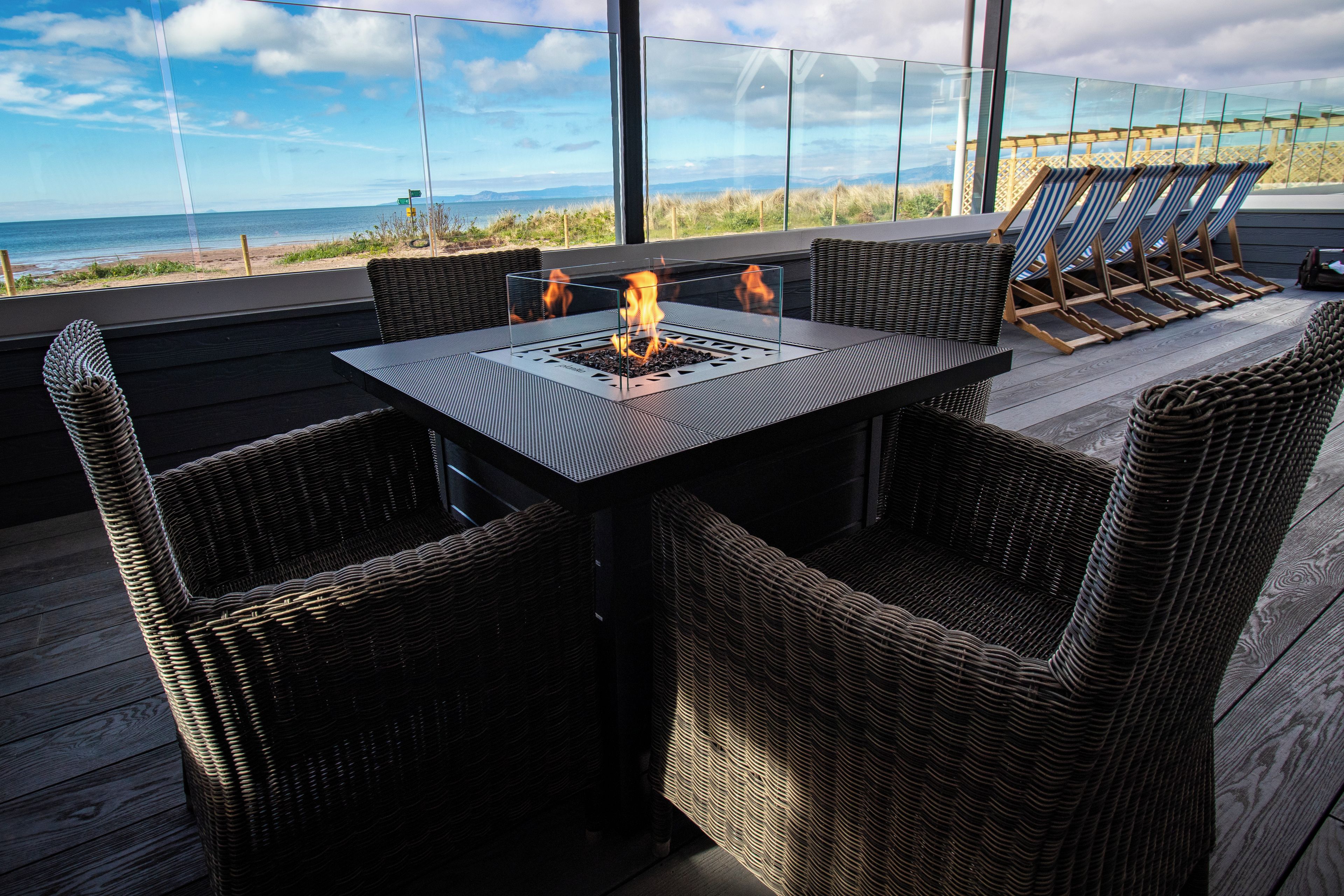 A deck with wicker furniture, a glass-top table, and a fire feature.