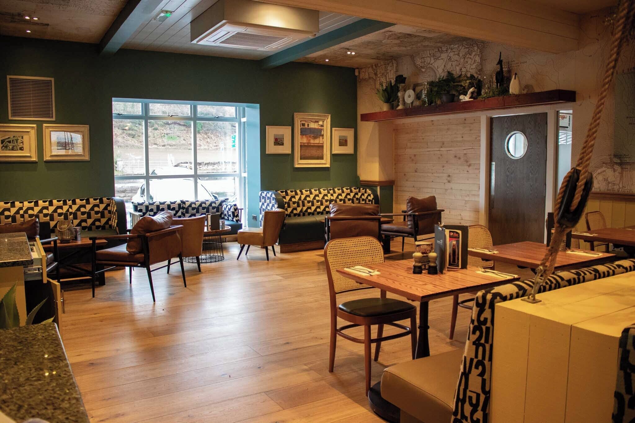 Un restaurante con suelo de madera, un ventanal grande y una zona de estar con cojines estampados.