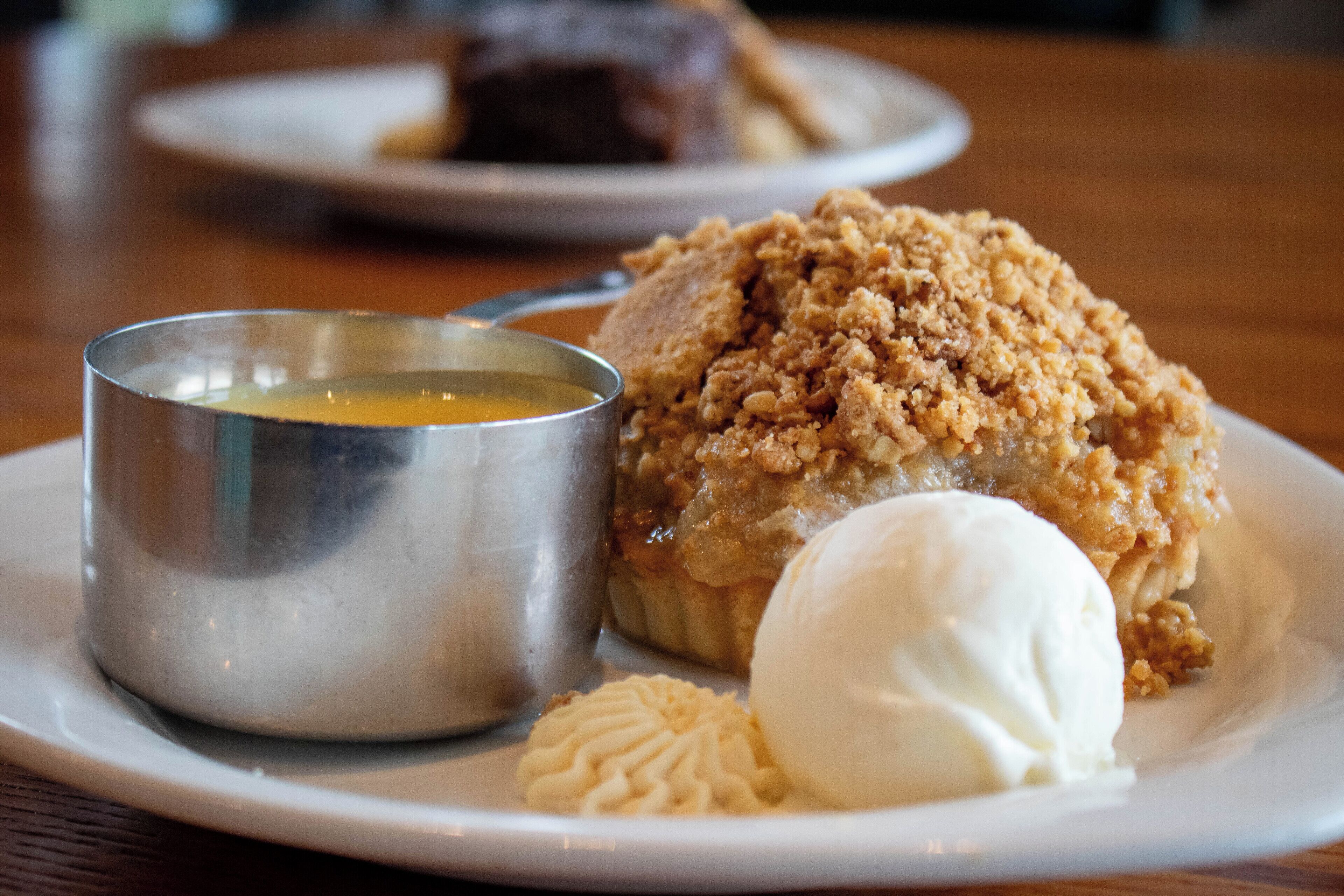 Un plato de postre con un dulce cubierto de crumble, una bola de helado y una pequeña taza de metal.