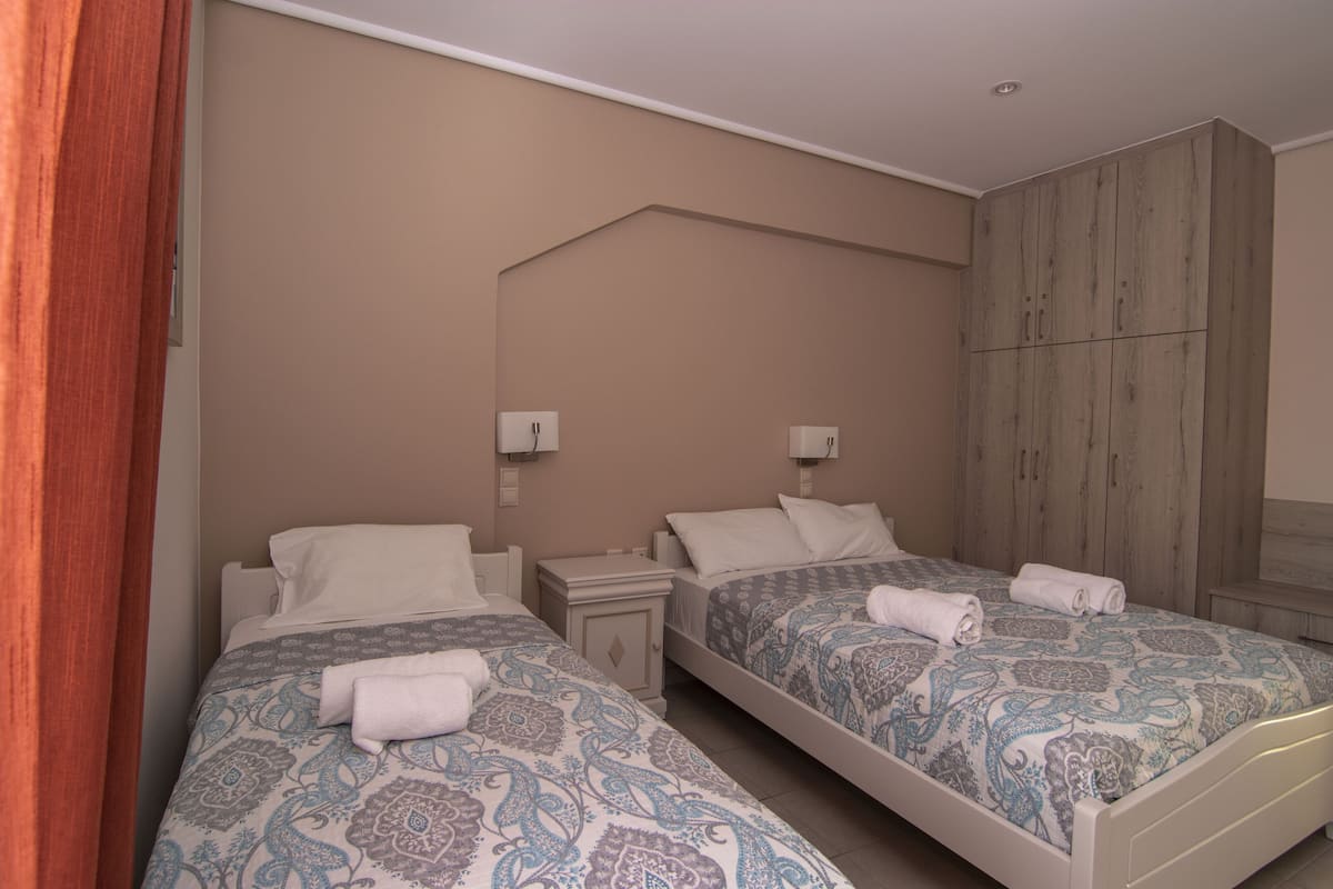 A hotel room with two beds, each with patterned bedding and towels. There are bedside tables with lamps and a wardrobe.