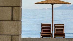 Two wooden chairs under a thatched umbrella by a body of water, with a stone pillar and a street lamp in the foreground. - Nefeli Studios (Dorida)
