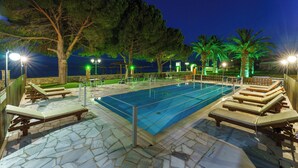 A swimming pool with lounge chairs and a stone deck, surrounded by trees and palm trees, with a view of the sea at night. - Nefeli Studios (Dorida)