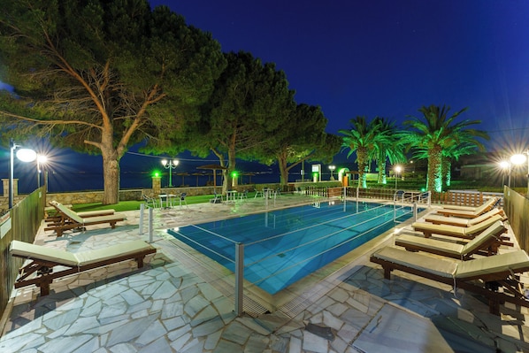 A swimming pool with lounge chairs and a stone deck, surrounded by trees and palm trees, with a view of the sea at night.