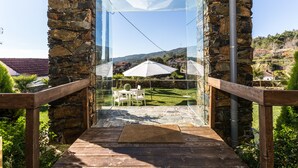 A glass-enclosed balcony with stone walls, a wooden deck, and a view of a grassy area with umbrellas. - A Casa do Tio Tenente (Serta)