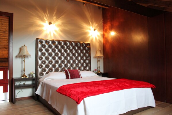 Una habitación de hotel con un cabecero capitonado, dos lámparas de noche, una manta roja y suelo de madera.