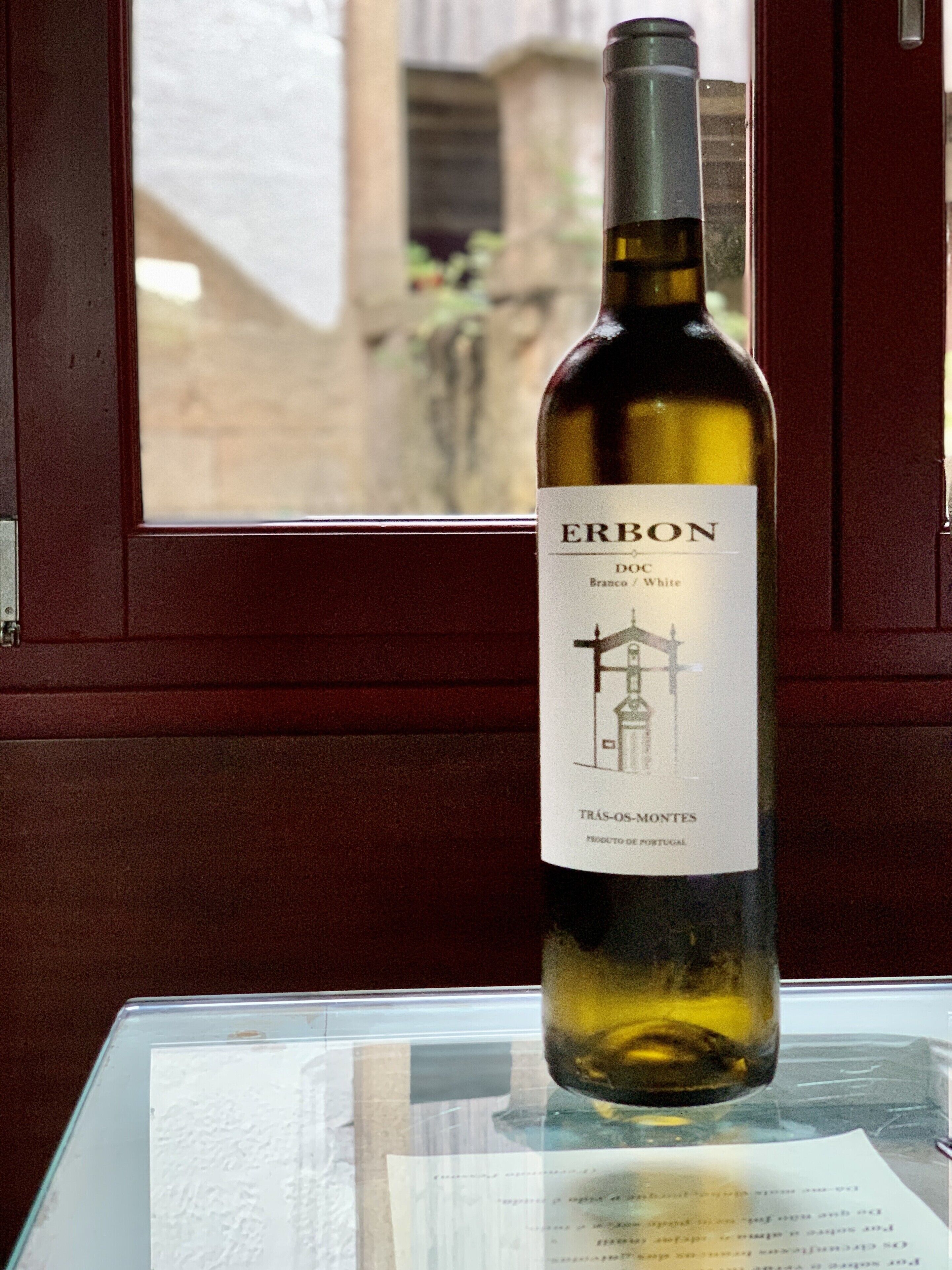 A bottle of Erbon DOC Branco/White wine with a label featuring a building illustration, placed on a table with a window in the background.
