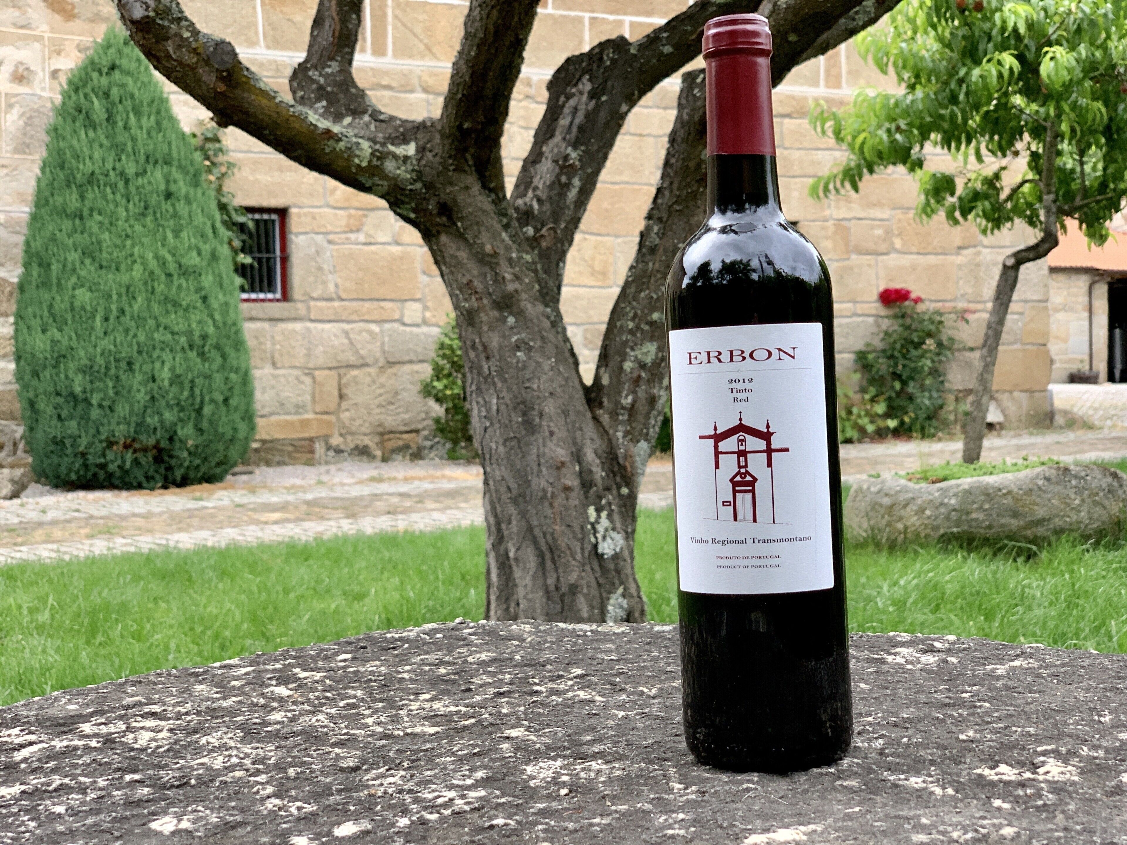 Una botella de vino Erbon sobre una superficie de piedra, con un edificio de piedra y un árbol al fondo.