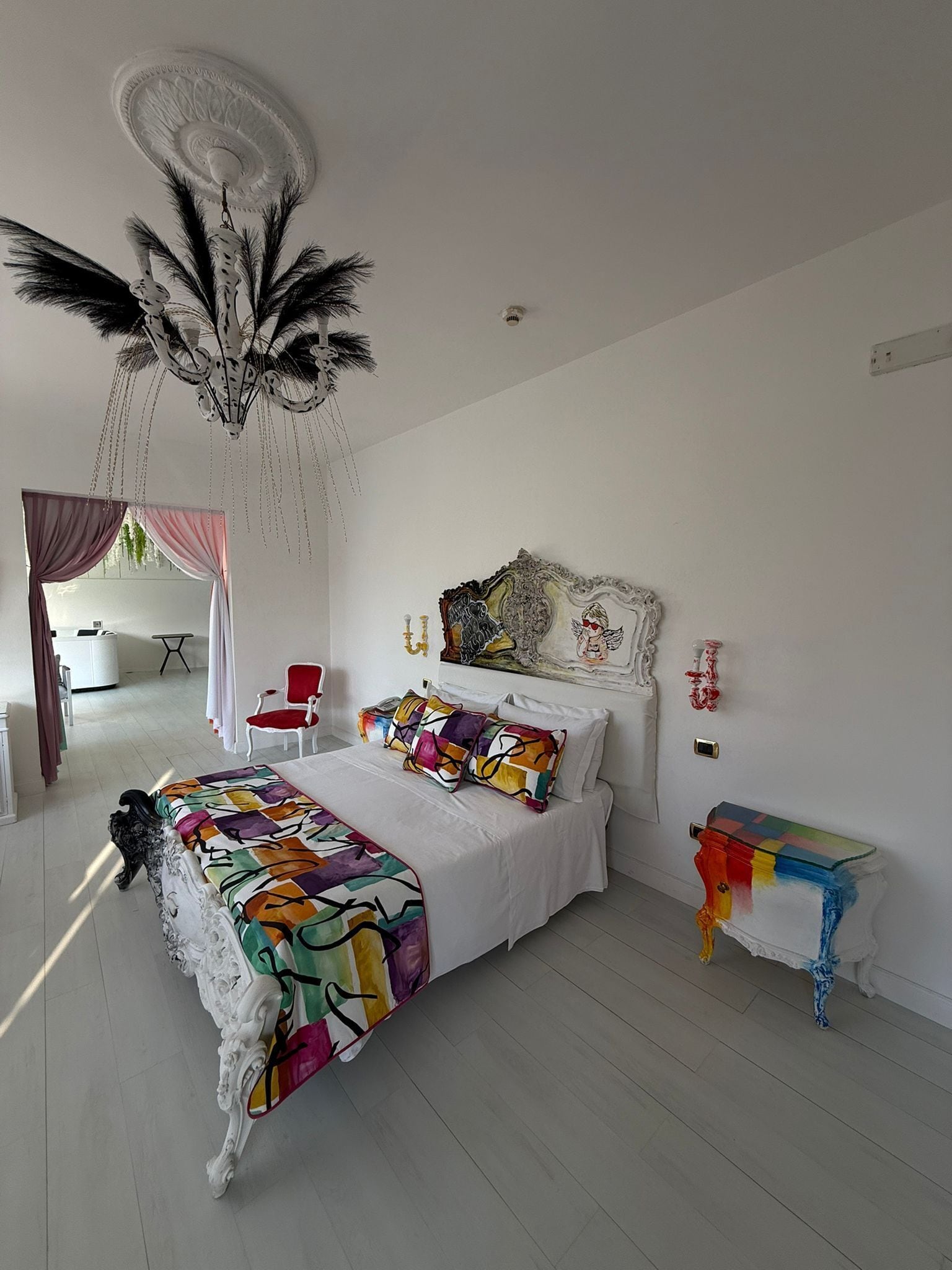 Ein Schlafzimmer mit einem großen Bett, einer Kandelaber, einem roten Sessel und einem farbenfrohen Beistelltisch.