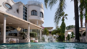 A pool area with a fountain, surrounded by palm trees and a building with a curved facade and large windows. - Hotel Imperiale (Terzigno)